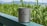 A stainless steel mug with the brand name 'snow peak' inscribed on it sits on a green railing. In the background, there is lush green foliage and some distant mountains under a clear blue sky.