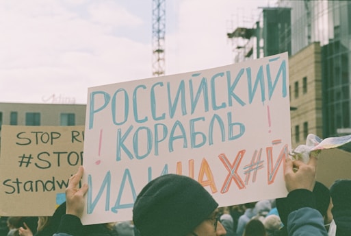 A protest scene with people holding signs in various languages. One sign features a message written in Russian with large blue and red letters. Another sign nearby includes the phrases 'STOP' and '#STANDWITH'. The background shows a cityscape with modern buildings and a crane.