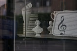 A display window featuring various music-themed items, including a bust of a historical figure, a folded paper with musical notes, and a tray adorned with sheet music and a large treble clef symbol. The objects are set against a blurred background of a street scene.