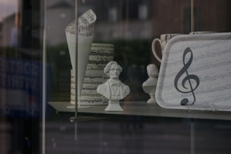 A display window featuring various music-themed items, including a bust of a historical figure, a folded paper with musical notes, and a tray adorned with sheet music and a large treble clef symbol. The objects are set against a blurred background of a street scene.