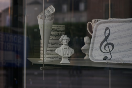 A display window featuring various music-themed items, including a bust of a historical figure, a folded paper with musical notes, and a tray adorned with sheet music and a large treble clef symbol. The objects are set against a blurred background of a street scene.