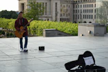 A musician playing an acoustic guitar plugged into a compact musiccube amp on a sunny park bench
