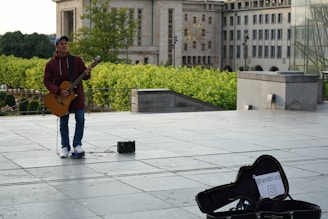 A street musician is playing an acoustic guitar in an urban plaza setting. He is wearing a maroon jacket and a blue cap, standing on a large paved area. There's a small amplifier on the ground near him, and an open guitar case with a sign inside featuring an Instagram handle. The background includes lush greenery and a large stone building.