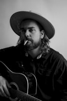 A person with long hair and a beard is wearing a wide-brimmed hat while holding an acoustic guitar. They have a cigarette in their mouth, and smoke is gently rising. The person wears a dark shirt, and the image is in black and white, giving it a classic, timeless feel.