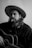 A person with long hair and a beard is wearing a wide-brimmed hat while holding an acoustic guitar. They have a cigarette in their mouth, and smoke is gently rising. The person wears a dark shirt, and the image is in black and white, giving it a classic, timeless feel.
