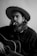 A person with long hair and a beard is wearing a wide-brimmed hat while holding an acoustic guitar. They have a cigarette in their mouth, and smoke is gently rising. The person wears a dark shirt, and the image is in black and white, giving it a classic, timeless feel.