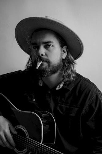 A person with long hair and a beard is wearing a wide-brimmed hat while holding an acoustic guitar. They have a cigarette in their mouth, and smoke is gently rising. The person wears a dark shirt, and the image is in black and white, giving it a classic, timeless feel.