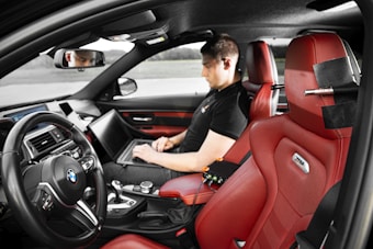 A person is sitting in the driver's seat of a car, working on a laptop placed on their lap. The car has red leather seats and a high-tech dashboard. The steering wheel prominently displays the BMW logo. The interior appears modern and sleek, with a focus on technology.
