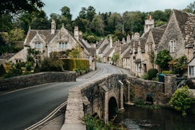 A quaint village scene featuring traditional stone cottages with steep roofs, lush greenery, and a narrow stone bridge crossing a small creek. The road curves gently through the village, lined with well-maintained hedges and flower gardens. The overall atmosphere is peaceful and timeless.