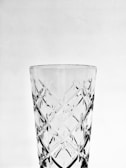 A transparent glass with intricate cut patterns is centered against a plain background. The surface features crisscross diamond shapes, enhancing its elegance and complexity.
