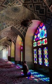 A person sitting inside a beautifully decorated mosque with intricate mosaic patterns on the ceiling. Vibrant stained glass windows cast colorful light patterns on the floor and walls, creating a serene and spiritual atmosphere.
