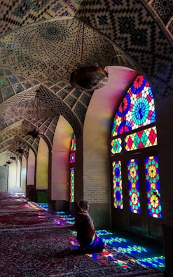 A person sitting inside a beautifully decorated mosque with intricate mosaic patterns on the ceiling. Vibrant stained glass windows cast colorful light patterns on the floor and walls, creating a serene and spiritual atmosphere.