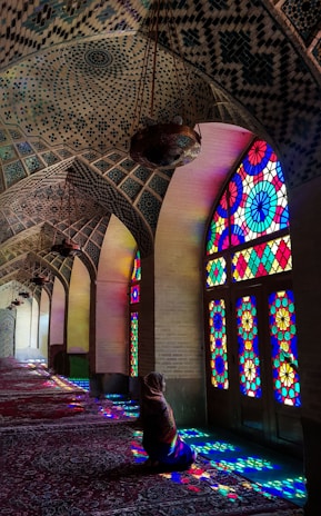 A person sitting inside a beautifully decorated mosque with intricate mosaic patterns on the ceiling. Vibrant stained glass windows cast colorful light patterns on the floor and walls, creating a serene and spiritual atmosphere.