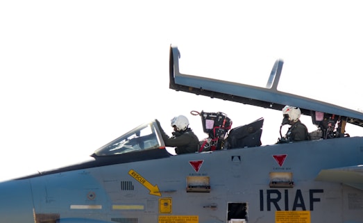 Two pilots wearing helmets are seated in the cockpit of a military aircraft. The cockpit canopy is open, and the aircraft has various markings, including the acronym 'IRIAF' on its body. The pilots are focused, suggesting a pre-flight preparation or taxiing scenario.
