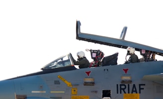Two pilots wearing helmets are seated in the cockpit of a military aircraft. The cockpit canopy is open, and the aircraft has various markings, including the acronym 'IRIAF' on its body. The pilots are focused, suggesting a pre-flight preparation or taxiing scenario.