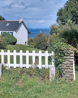 A scenic view featuring a white picket fence in the foreground, lush greenery, and a small house with a dark roof. In the background, the ocean is visible with a couple of sailboats on the horizon under a partly cloudy sky.