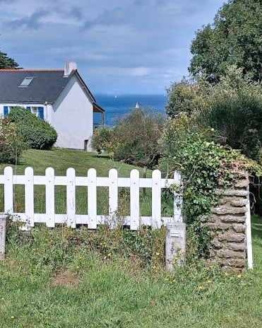 A scenic view featuring a white picket fence in the foreground, lush greenery, and a small house with a dark roof. In the background, the ocean is visible with a couple of sailboats on the horizon under a partly cloudy sky.