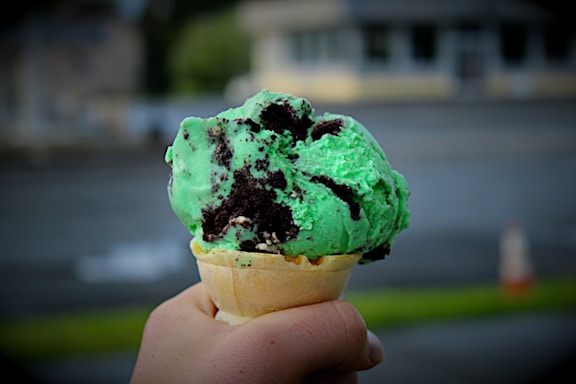 A hand holding an ice cream cone filled with green mint chocolate chip ice cream. The ice cream is packed with chocolate chunks and is set against an out-of-focus urban background.