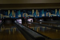A dimly lit bowling alley with multiple lanes featuring a vibrant mural of cityscape art above the pins. Several bowling pins are set up in their positions with a green bowling ball rolling towards them on lane number 5.