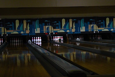 A dimly lit bowling alley with multiple lanes featuring a vibrant mural of cityscape art above the pins. Several bowling pins are set up in their positions with a green bowling ball rolling towards them on lane number 5.
