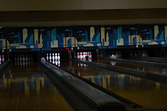 A dimly lit bowling alley with multiple lanes featuring a vibrant mural of cityscape art above the pins. Several bowling pins are set up in their positions with a green bowling ball rolling towards them on lane number 5.
