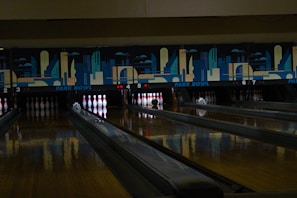 A dimly lit bowling alley with multiple lanes featuring a vibrant mural of cityscape art above the pins. Several bowling pins are set up in their positions with a green bowling ball rolling towards them on lane number 5.