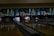 A dimly lit bowling alley with multiple lanes featuring a vibrant mural of cityscape art above the pins. Several bowling pins are set up in their positions with a green bowling ball rolling towards them on lane number 5.