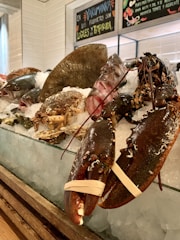 A vibrant seafood display featuring a variety of fresh shellfish and fish laid out on a bed of ice. There are lobsters with rubber bands around their claws, crabs, oysters, and several types of fish. Behind the display, chalkboard signs with colorful writing advertise the freshness and local sourcing of the produce.