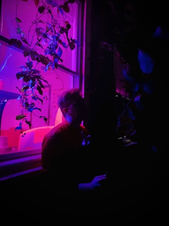 A warm, softly lit portrait of Luna seated by a window framed with leafy plants, bathed in gentle purple tones.