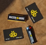 Several items are arranged on a brown textured surface, including a black and yellow gift card, a lighter with branding, and a box labeled 'Buzzed Buds'. The logo features a stylized bee symbol.