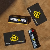 Several items are arranged on a brown textured surface, including a black and yellow gift card, a lighter with branding, and a box labeled 'Buzzed Buds'. The logo features a stylized bee symbol.