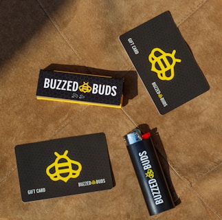 Several items are arranged on a brown textured surface, including a black and yellow gift card, a lighter with branding, and a box labeled 'Buzzed Buds'. The logo features a stylized bee symbol.