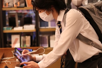 A person wearing a white shirt, tie, and surgical mask uses a stylus on a tablet device. They have a gray backpack and are in an interior space with shelves filled with books or similar items in the background.