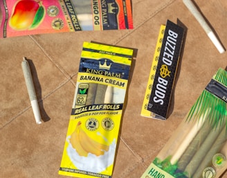 Several packages of flavored leaf rolls are displayed on a brown surface. The packets include 'Banana Cream' and feature vibrant colors with fruits depicted. There are also pre-rolled cones on the surface.