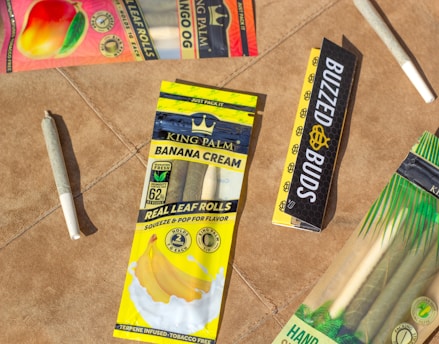 Several packages of flavored leaf rolls are displayed on a brown surface. The packets include 'Banana Cream' and feature vibrant colors with fruits depicted. There are also pre-rolled cones on the surface.
