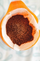 A close-up view of a paper filter containing freshly ground coffee, placed in the top section of a coffee maker. The filter sits atop an orange container, with a focus on the texture and rich brown color of the coffee grounds.
