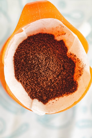 A close-up view of a paper filter containing freshly ground coffee, placed in the top section of a coffee maker. The filter sits atop an orange container, with a focus on the texture and rich brown color of the coffee grounds.