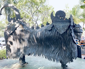A large, intricately designed metal sculpture of a mythical creature with a lion-like body, thick mane, and decorative patterns on its back. The sculpture is set outdoors in a park-like area surrounded by trees and greenery.