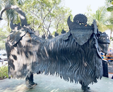 A large, intricately designed metal sculpture of a mythical creature with a lion-like body, thick mane, and decorative patterns on its back. The sculpture is set outdoors in a park-like area surrounded by trees and greenery.
