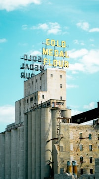 An industrial building with multiple concrete silos and a prominent yellow sign on the rooftop that reads 'Gold Medal Flour.' The building appears weathered and historical, with a few small windows and brickwork at the lower levels. The sky is a bright and clear blue with scattered clouds.