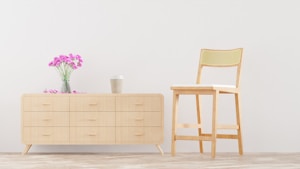 A minimalist room with a light wood dresser on the left adorned with a vase of pink flowers. A tall chair with a light fabric cushion is placed on the right. The wall and floor are in neutral tones, enhancing the clean and simple aesthetic.