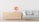 A minimalist interior features a light-colored wooden dresser with six drawers on the left side and a light grey sofa with four cushions on the right. There is a vase with red flowers placed on top of the dresser. The background wall is a plain white, and the floor has a light wooden texture.