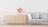 A minimalist interior features a light-colored wooden dresser with six drawers on the left side and a light grey sofa with four cushions on the right. There is a vase with red flowers placed on top of the dresser. The background wall is a plain white, and the floor has a light wooden texture.