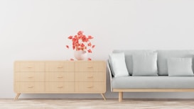 A minimalist interior features a light-colored wooden dresser with six drawers on the left side and a light grey sofa with four cushions on the right. There is a vase with red flowers placed on top of the dresser. The background wall is a plain white, and the floor has a light wooden texture.