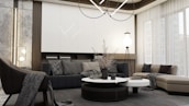 A modern living room with a minimalist design features a large sectional sofa in neutral tones, complemented by assorted cushions with subtle patterns. A round coffee table with decorative items sits in the center. The room is illuminated by a contemporary chandelier and additional lighting fixtures. Floor-to-ceiling windows with sheer curtains allow natural light to flood in, enhancing the spacious and serene atmosphere.