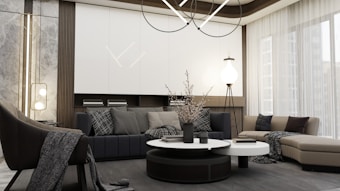 A modern living room with a minimalist design features a large sectional sofa in neutral tones, complemented by assorted cushions with subtle patterns. A round coffee table with decorative items sits in the center. The room is illuminated by a contemporary chandelier and additional lighting fixtures. Floor-to-ceiling windows with sheer curtains allow natural light to flood in, enhancing the spacious and serene atmosphere.
