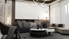 A modern living room with a minimalist design features a large sectional sofa in neutral tones, complemented by assorted cushions with subtle patterns. A round coffee table with decorative items sits in the center. The room is illuminated by a contemporary chandelier and additional lighting fixtures. Floor-to-ceiling windows with sheer curtains allow natural light to flood in, enhancing the spacious and serene atmosphere.