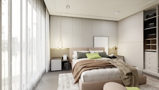 A modern bedroom features a large bed with a variety of pillows and throws in neutral tones, complemented by a soft area rug. A pendant light hangs to one side, and built-in storage cupboards are visible on the opposite wall. Large sheer curtains allow for ample natural light to brighten the space.