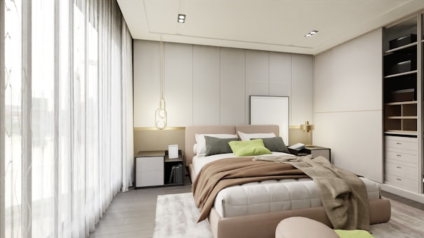 A modern bedroom features a large bed with a variety of pillows and throws in neutral tones, complemented by a soft area rug. A pendant light hangs to one side, and built-in storage cupboards are visible on the opposite wall. Large sheer curtains allow for ample natural light to brighten the space.