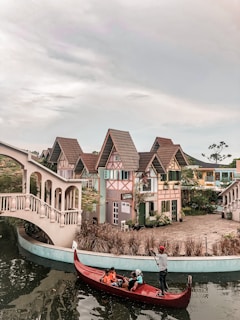 A picturesque scene featuring a quaint village with charming, colorful houses sporting steep, tiled roofs reminiscent of European architecture. A canal runs in the foreground, spanned by an elegant, arched bridge. A person navigates a red gondola on the water, carrying several passengers, adding a touch of Venetian charm to the setting. The area is lush with greenery, complementing the whimsical feel of the village.